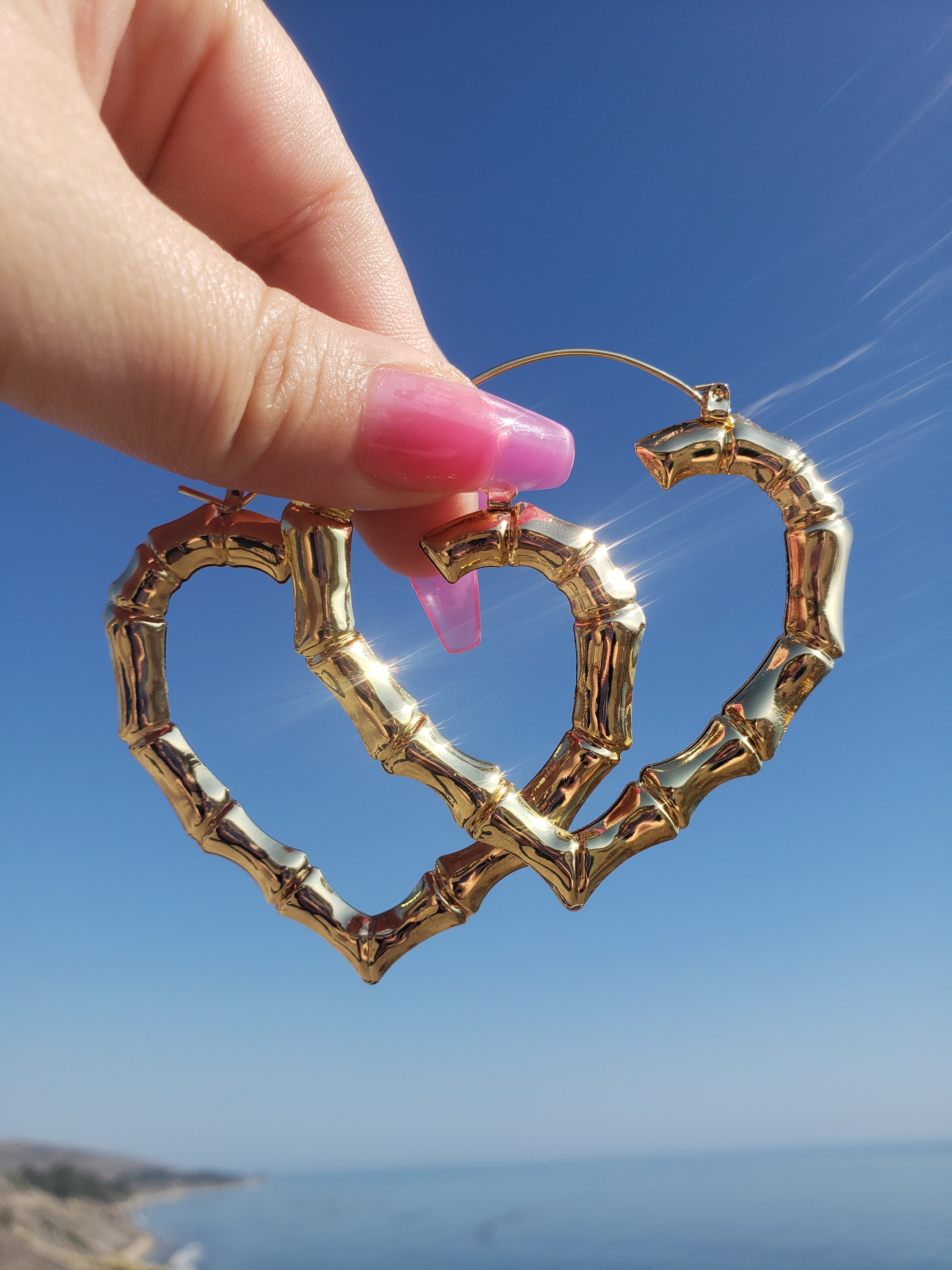 Women's hand with Pink long nails holding Super Light & Thin Gold Plated Heart Bamboo Hoop Earrings. Background at a beach scenery. most of the picture is a beautiful blue gradient sky with a slight view of the beautiful blue ocean and an even slighter view  on the left size corner of the California 101 freeway coastline. 