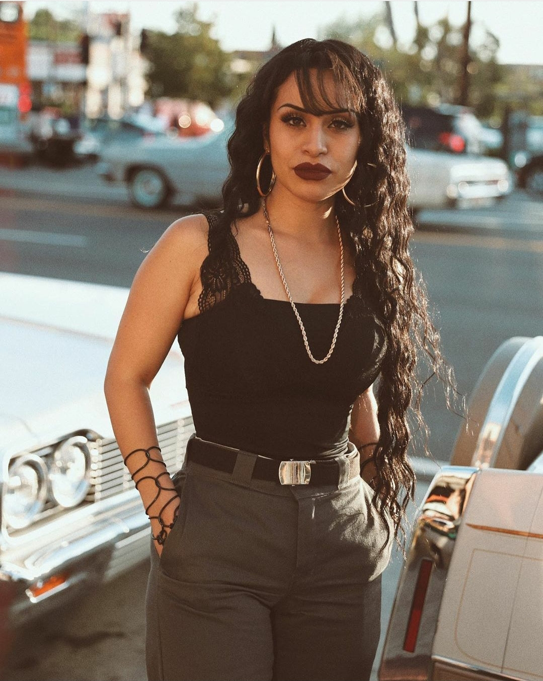 Brown indigenous chicana women dressed in a classic chola look. Dickies dark gray pants with basic black belt and black cami. She is wearing our big glam hoops as well as our 26inches twisted chain necklaces. Her hair is down scunched curly but down. Her hair falls down to her hips and has some selena styled bangs. Background san diego streets you can see creamed colored lowrider cars in the background. 