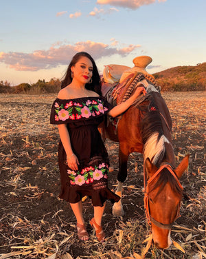 Photography of a model next to a horse. Mexicana wearing a black off the shoulder dress  with beautiful traditional mexican flower embroidery design on the top ruffle and bottom with lace trip details in between the bottom embroidery. Flowers are pink and red colored. Model is next to brown horse in a corn field rancho. The dress is for sale, made in mexico